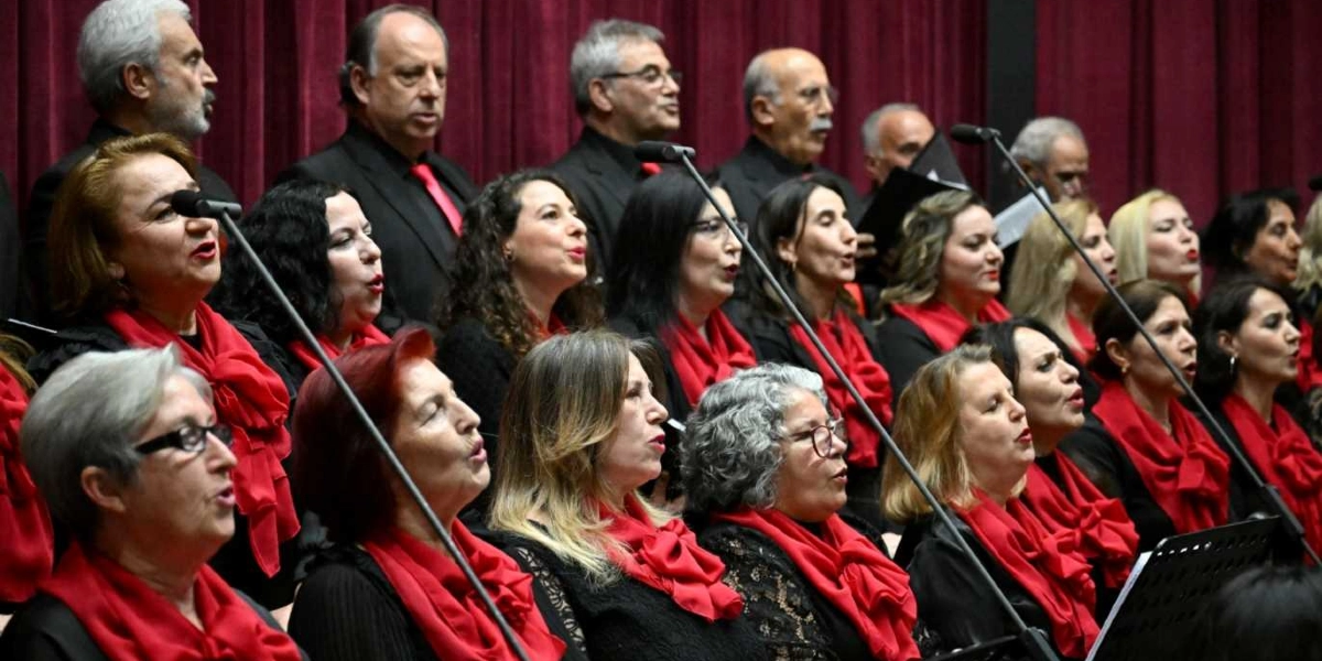 Siyah kıyafetler giymiş, boyunlarında kırmızı fularlar bulunan kadın ve erkeklerden oluşan bir koro, bordo renkli perde önünde mikrofonlar eşliğinde şarkı söylüyor.