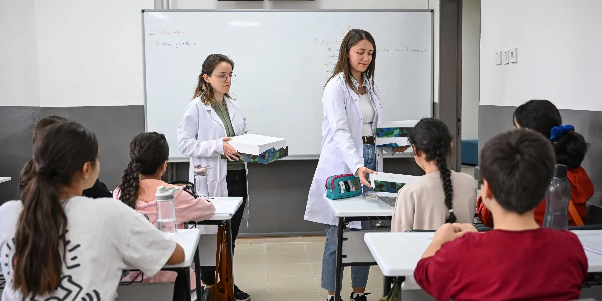 Beyaz önlüklü iki eğitmen, bir sınıf ortamında öğrencilere üzerinde geometrik desenler bulunan beyaz kutular dağıtıyor. Öğrenciler sıralarında otururken, eğitmenlerden biri bir öğrenciye kutusunu uzatıyor, diğeri ise sınıfın ön tarafında elinde kutuyla bekliyor. Arka planda üzerinde ders notları yazılı olan beyaz bir yazı tahtası bulunuyor.