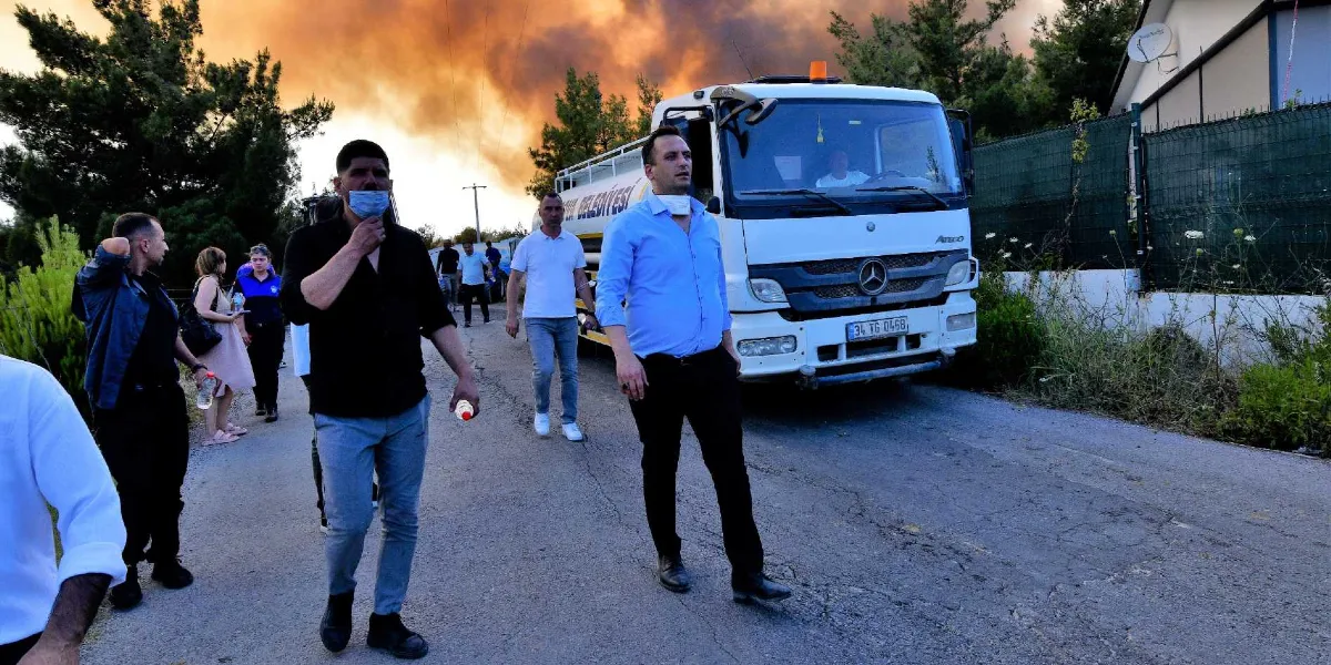 Yakaköy'de orman yangınının neden olduğu yoğun duman ve turuncu gökyüzü altında, asfalt bir yolda yürüyen bir grup insan ve yanlarında duran bir belediye su tankeri. Ön planda siyah gömlekli ve açık mavi gömlekli iki erkek yürürken, arka planda diğer vatandaşlar ve ağaçlık alanın üzerinde yükselen yangın dumanları görülüyor.