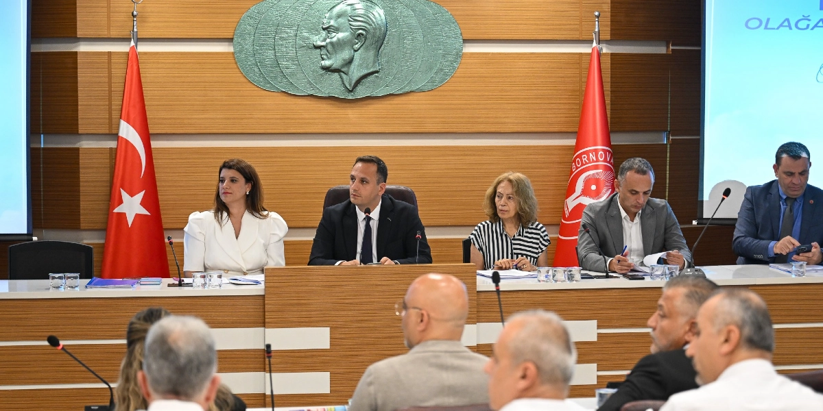 Bornova Belediyesi'ne ait bir toplantı salonunda, ahşap kaplama duvar önünde Türk bayrağı ve Bornova Belediyesi flaması arasında oturan beş kişilik bir heyet yer alıyor. Heyetin arkasındaki duvarda Atatürk'ün kabartma portresi bulunuyor. Masada oturanlar arasında beyaz ceketli bir kadın, takım elbiseli erkekler ve çizgili bluzlu bir kadın bulunuyor; önlerinde mikrofonlar ve belgeler yer alıyor. Toplantı salonunun ön kısmında ise katılımcıların sırtları dönük şekilde oturduğu görülüyor.