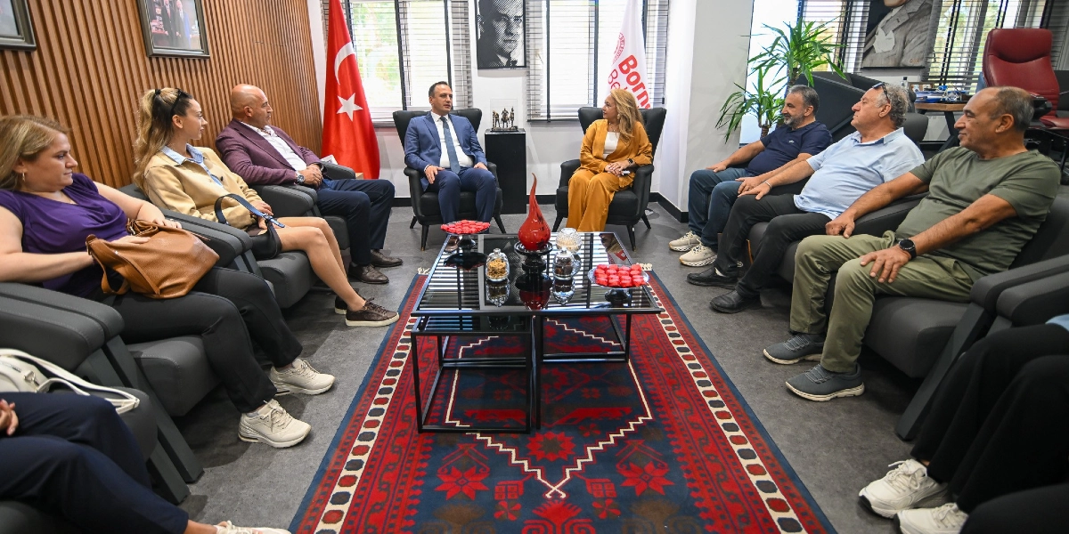 Modern bir ofis ortamında, karşılıklı siyah koltuklarda oturan bir grup insan toplantı yapıyor. Odanın merkezinde, üzerinde kırmızı süslemeler ve ikramlar bulunan cam bir sehpa yer alıyor. Arka planda Türk bayrağı, Bornova Belediyesi flaması ve duvarda Atatürk portresi bulunuyor. Takım elbiseli bir erkek ve sarı kıyafetli bir kadın, grubun merkezinde yer alarak toplantıyı yönetiyor. Diğer katılımcılar günlük kıyafetleriyle onları dinliyor.