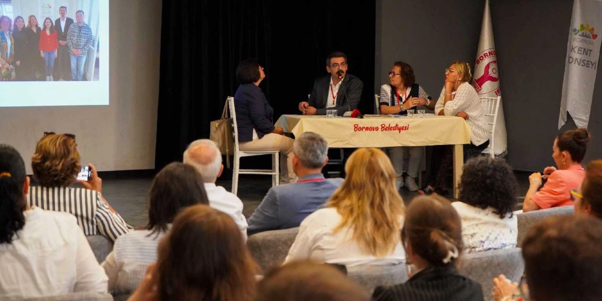 Bornova Kent Konseyi toplantısında, üzerinde Bornova Belediyesi yazılı bir masanın etrafında oturan üç konuşmacı ve onları dinleyen katılımcılar yer alıyor. Sahnedeki konuşmacıların arkasındaki duvarda, bir grup insanın yer aldığı bir fotoğrafın yansıtıldığı projeksiyon perdesi ve yanında Bornova Kent Konseyi bayrağı bulunuyor. Katılımcılar salonun düzenine uygun şekilde oturarak konuşmayı takip ediyor.