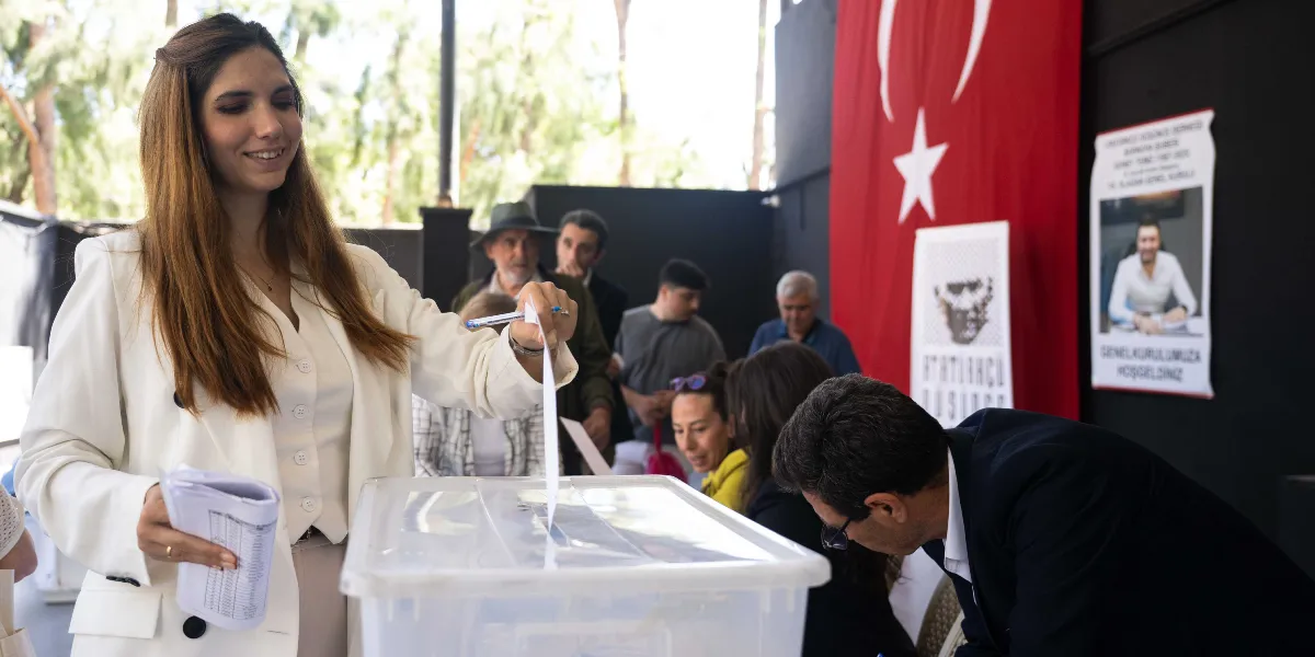 Atatürkçü Düşünce Derneği Genel Kurulu'nda, beyaz ceketli genç bir kadın elindeki oy pusulasını şeffaf sandığa atıyor. Arka planda Türk bayrağı, üzerinde Atatürk portresi bulunan bir afiş ve Güney Temiz'in fotoğrafının yer aldığı 