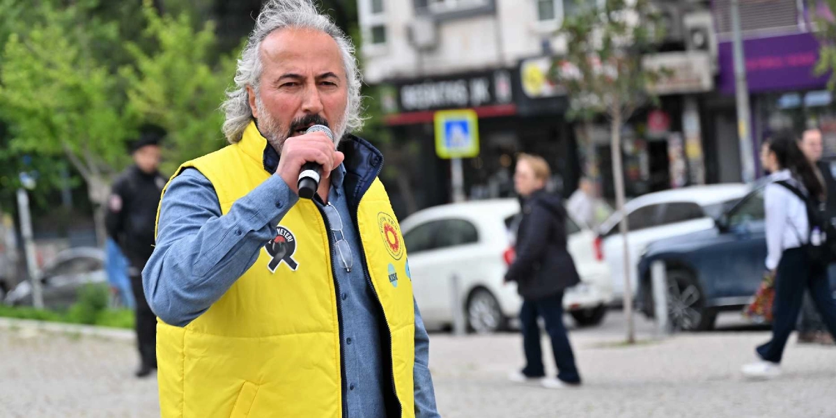 Gri saçlı ve sakallı bir erkek, üzerinde Eğitim Sen logosu bulunan sarı bir yelek giymiş şekilde elinde mikrofonla konuşma yapıyor. Arka planda şehir merkezinde bir cadde, araçlar ve yoldan geçen insanlar görülüyor.