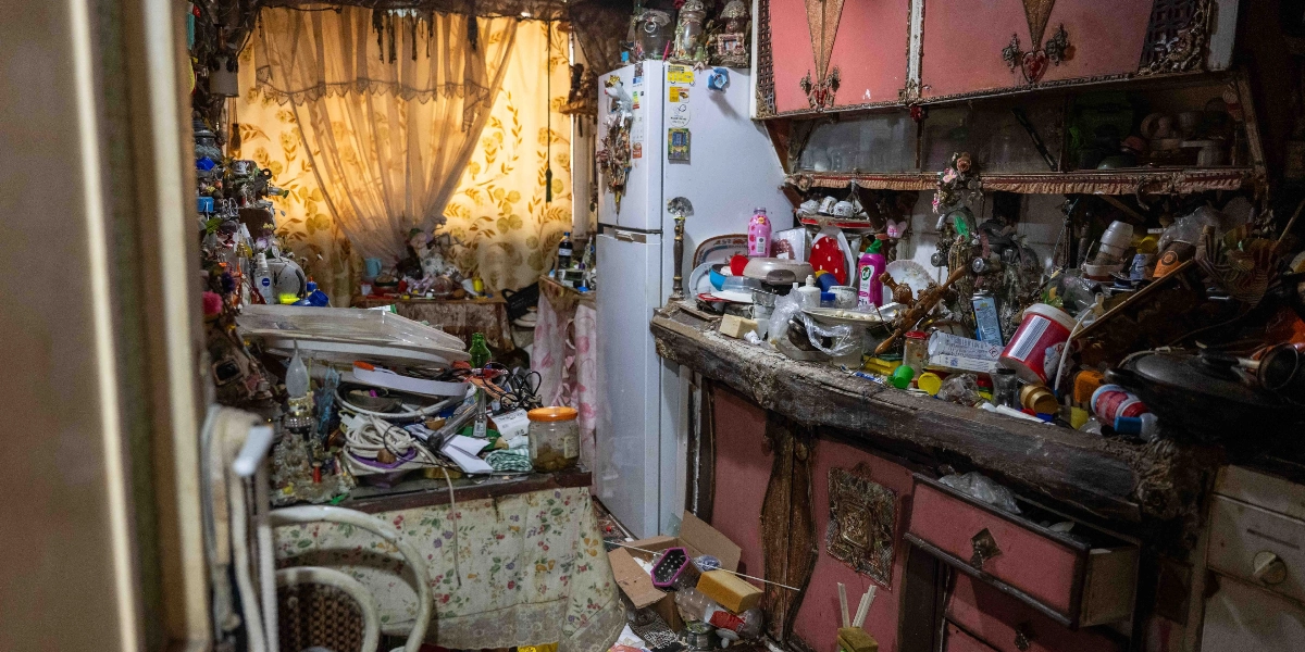 Eski ve yıpranmış mutfak dolaplarıyla dolu, tezgahı ve masası çok sayıda mutfak eşyası, plastik atık, şişe ve çeşitli ev eşyalarıyla tamamen kaplanmış düzensiz bir mutfak. Beyaz bir buzdolabının önü ve çevresi de dahil olmak üzere her yerin eşyalarla yığılı olduğu, pencerelerin önünde ise çiçek desenli perdelerin asılı olduğu dağınık bir yaşam alanı.