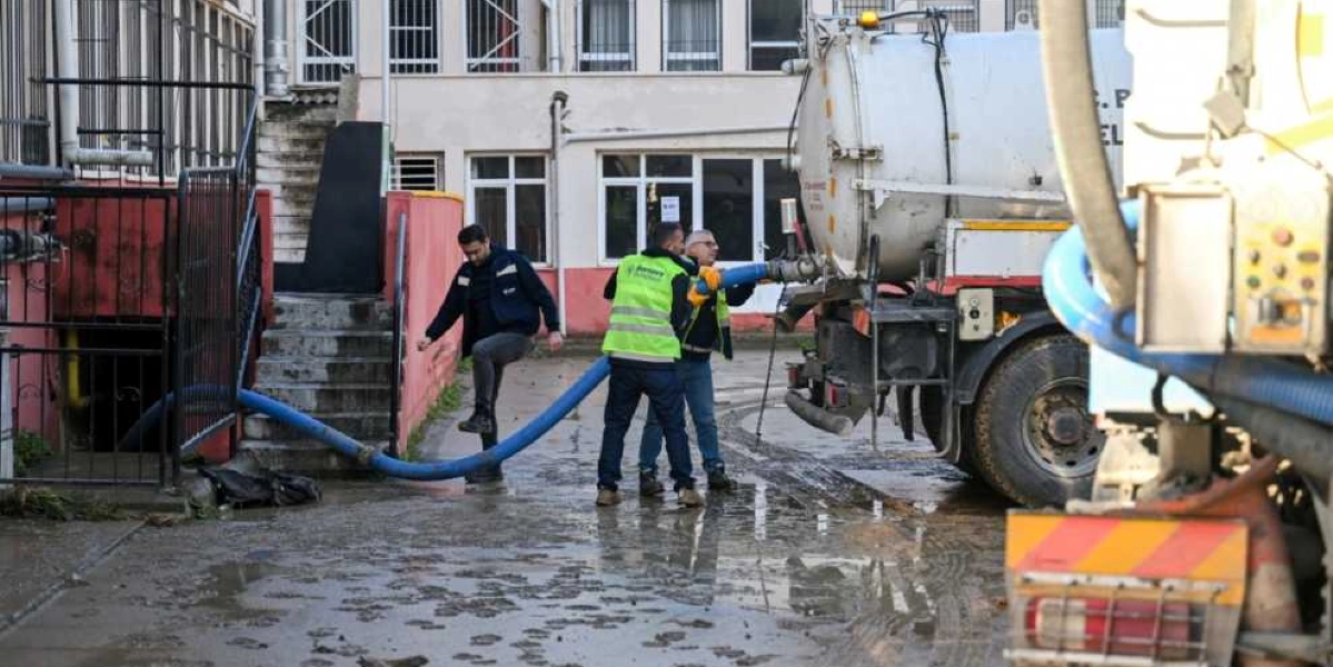 Bornova Belediyesi personeli, sel sonrası çamurlu bir bahçede vidanjör aracı kullanarak su tahliye çalışması yapıyor. Üzerinde Bornova Belediyesi logolu fosforlu yelek bulunan bir görevli hortumu tutarken, diğer iki görevli çalışmaya destek veriyor.