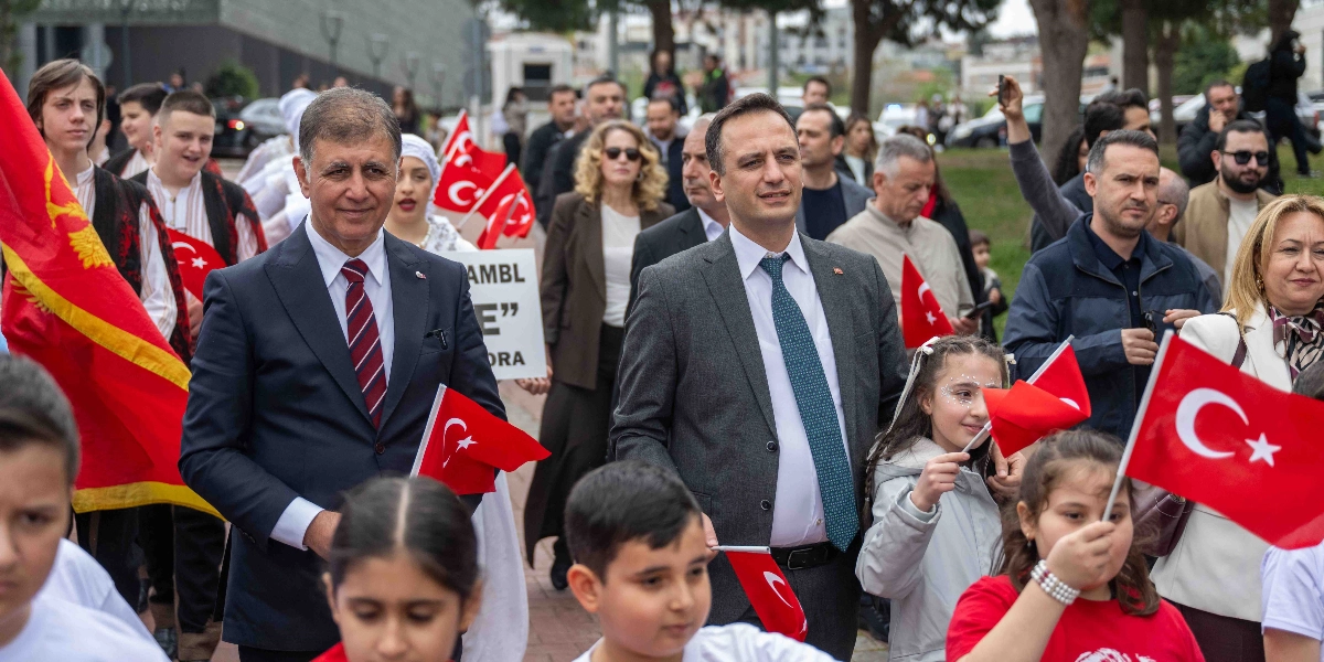 Bornova'da düzenlenen bir etkinlikte, ellerinde Türk bayrakları taşıyan çocuklar ve onlara eşlik eden yetişkinlerin yer aldığı bir yürüyüş korteji. Ön planda takım elbiseli iki erkek, arkalarında ise geleneksel kıyafetler giymiş gençler ve ellerinde bayraklarla yürüyen çocuklar bulunuyor. Arka planda ise ağaçlıklı bir yol ve etkinliği takip eden diğer katılımcılar görülüyor.