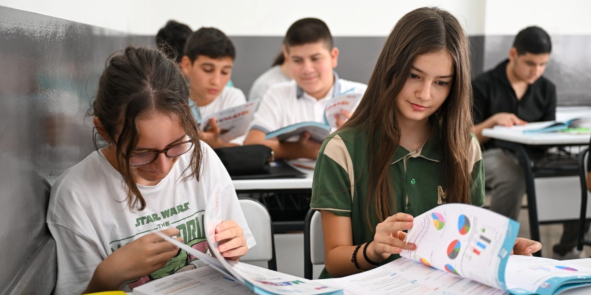 Sınıf ortamında ders çalışan öğrenciler, önlerinde açık kitaplar ve grafikli çalışma kağıtlarıyla masalarında oturuyor. Ön planda beyaz tişörtlü gözlüklü bir kız öğrenci ile yeşil yakalı tişört giyen uzun saçlı bir kız öğrenci ders materyallerini inceliyor; arka planda ise diğer öğrenciler kitaplarını okumaya devam ediyor.