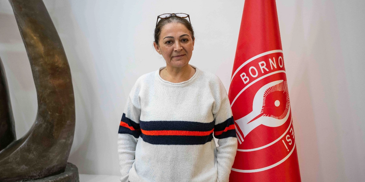 Koyu renkli saçlarını arkadan toplamış, başında gözlük olan, üzerinde beyaz üzerine kırmızı ve lacivert çizgili bir kazak bulunan orta yaşlı bir kadın, yanında Bornova Belediyesi logosunun yer aldığı kırmızı bir bayrakla poz veriyor.