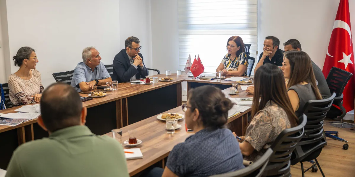 Aydınlık bir ofis ortamında, U şeklinde birleştirilmiş masaların etrafında toplanmış yaklaşık on kişilik bir grup toplantı yapıyor. Masanın üzerinde çay bardakları, tabaklar ve bazı belgeler bulunuyor. Katılımcılar dikkatle birbirlerini dinliyor, masanın başında ise Türk bayrağı asılı duruyor.