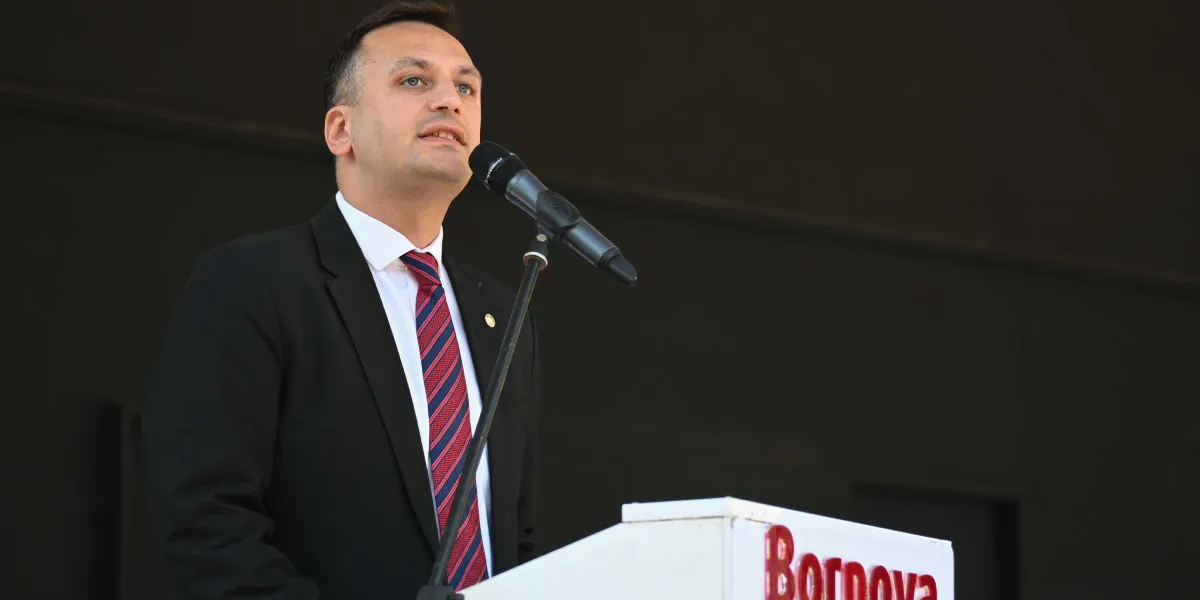 Koyu renkli bir arka plan önünde, beyaz gömlek, siyah ceket ve kırmızı-lacivert çizgili kravat giymiş bir erkek, üzerinde kırmızı renkle Bornova yazan beyaz bir kürsüde mikrofon başında konuşma yapıyor.
