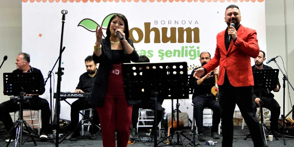 Bornova Tohum Takas Şenliği yazılı bir pankartın önünde sahne alan müzik grubu, mikrofonlarla şarkı söylüyor. Grubun önünde siyah kıyafetli ve bordo pantolonlu bir kadın solist ile kırmızı ceketli bir erkek solist bulunuyor. Arka planda ise klavye, bağlama ve diğer enstrümanları çalan müzisyenler yer alıyor.