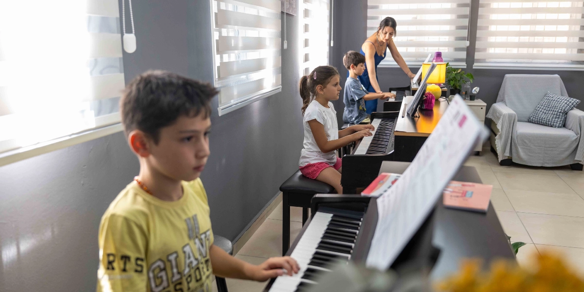Aydınlık bir sınıfta piyano başında oturan üç çocuk ve onlara rehberlik eden bir eğitmen. Ön planda sarı tişörtlü bir erkek çocuk, orta planda beyaz tişörtlü bir kız çocuk ve arka planda gri tişörtlü bir erkek çocuk piyanolarının başında oturuyor. Mavi elbiseli kadın eğitmen, çocuklara piyano çalarken eşlik ediyor. Odanın köşesinde gri bir koltuk ve pencerelerde stor perdeler bulunuyor.