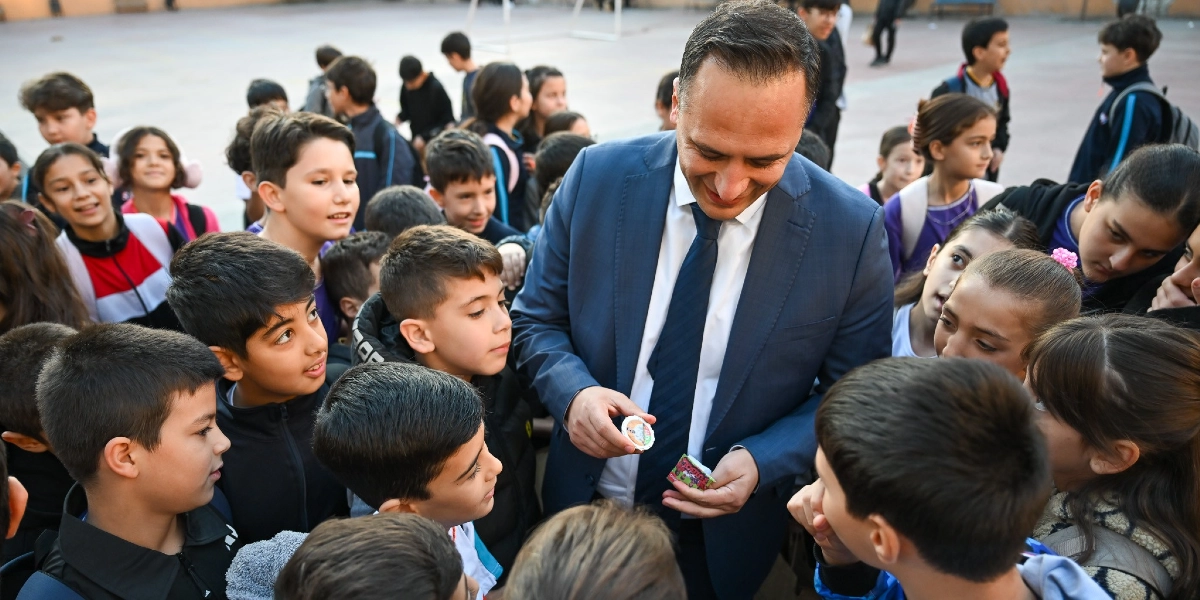 Lacivert takım elbiseli ve kravatlı bir erkek, okul bahçesinde etrafını saran ilkokul çağındaki öğrencilere küçük paketli yiyecekler dağıtıyor. Öğrenciler meraklı ve gülümseyen ifadelerle adama bakarken, adam elindeki beslenme paketlerini çocuklara uzatıyor.
