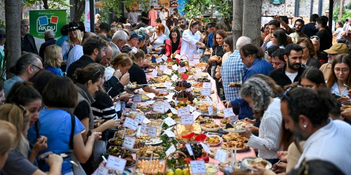 Açık havada kurulan uzun bir masanın etrafında toplanmış kalabalık bir grup insan, yerel yemeklerin ve ürünlerin sergilendiği bir tadım etkinliğine katılıyor. Masanın üzerinde çeşitli tabaklarda sunulan yiyecekler, meyveler ve her tabağın önünde ürünlerin isimlerinin yazılı olduğu küçük bilgi kartları yer alıyor. Arka planda ağaçlar ve taş duvarlı bir mekan görülürken, sol tarafta 