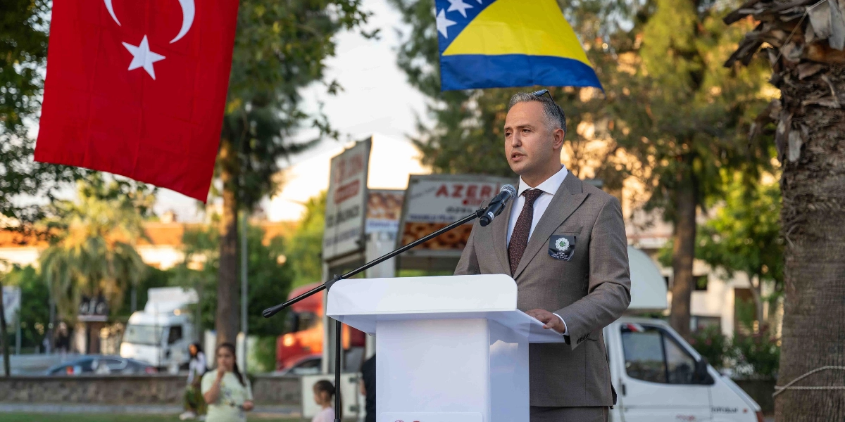 Açık havada, arkasında Türk bayrağı ve Bosna-Hersek bayrağı bulunan beyaz bir kürsüde konuşma yapan takım elbiseli bir erkek. Konuşmacının ceketinin sol yakasında Srebrenica soykırımını simgeleyen 