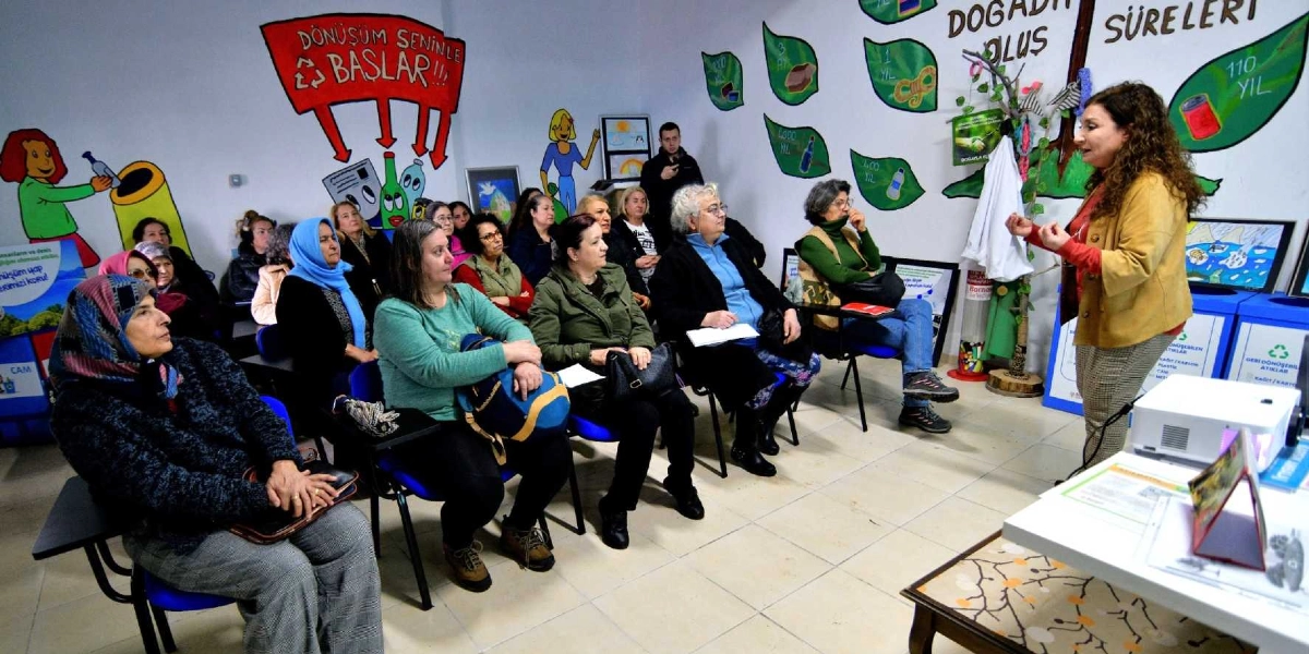 Eğitim amaçlı dekore edilmiş bir sınıfta, ayakta duran bir eğitmen, çoğunluğu kadınlardan oluşan katılımcı grubuna geri dönüşüm ve kompost üzerine sunum yapıyor. Duvarlarda 