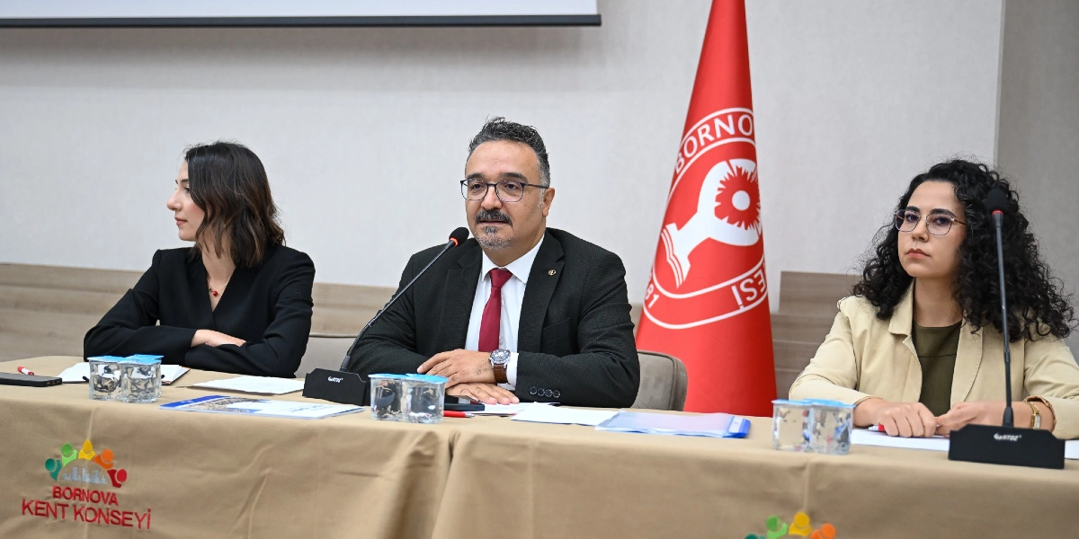 Bornova Kent Konseyi toplantısında, masanın arkasında yan yana oturan üç kişi yer alıyor. Ortada gözlüklü, koyu renk ceketli ve kırmızı kravatlı bir erkek konuşmacı bulunuyor. Konuşmacının sağında siyah ceketli bir kadın, solunda ise bej ceketli ve kıvırcık saçlı bir kadın oturuyor. Masanın üzerinde mikrofonlar, su bardakları ve bazı evraklar yer alıyor. Arka planda ise üzerinde Bornova Belediyesi logosu bulunan kırmızı bir bayrak duruyor. Masanın ön kısmında Bornova Kent Konseyi logosu görülüyor.