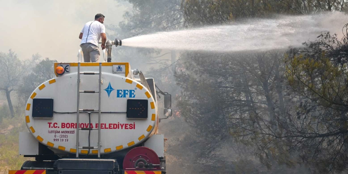 T.C. Bornova Belediyesi’ne ait bir su tankeri, ormanlık alandaki yangına müdahale ediyor. Tankerin üzerinde bulunan bir görevli, aracın üstündeki hortum düzeneğini kullanarak ağaçlara tazyikli su sıkıyor. Tankerin arkasında 