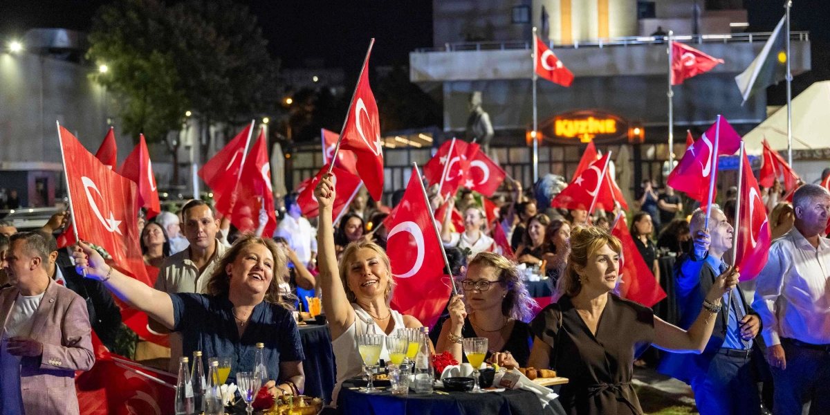 Gece vakti açık havada düzenlenen resepsiyonda, ellerinde Türk bayrakları tutan ve gülümseyerek kutlama yapan kalabalık bir grup insan. Ön planda masaların etrafında toplanmış, bayrak sallayan kadınlar ve arka planda ayakta duran katılımcılar yer alıyor. Arka planda ise ışıklandırılmış bir bina ve direklerde dalgalanan Türk bayrakları görülüyor.