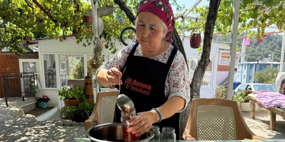 Başında kırmızı bir yazma bulunan, üzerinde Bornova Belediyesi logolu siyah bir önlük olan orta yaşlı bir kadın, dış mekanda bir masanın başında tencereden kepçeyle aldığı reçeli cam kavanoza dolduruyor. Arka planda asmalarla kaplı bir çardak, beyaz bir yapı ve park halindeki bir otomobil görülüyor.