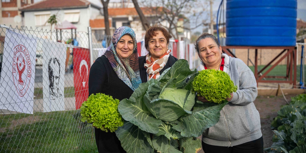 Bornova Kent Bostanı'nda hasat yapan üç kadın, ellerinde tuttukları büyük bir lahana ve marullarla kameraya gülümseyerek poz veriyor. Arka planda Bornova Belediyesi logosu, Atatürk silüeti ve Türk bayrağının bulunduğu bir çit ile büyük mavi bir su deposu yer alıyor.