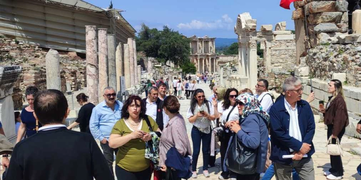Efes Antik Kenti'nde yürüyen, aralarında Bornova Muhtarlar Akademisi katılımcılarının da bulunduğu bir grup insan. Arka planda Celsus Kütüphanesi'nin tarihi sütunlu yapısı ve antik kalıntılar görülüyor. Gün ışığında, açık havada gerçekleşen gezide katılımcılar sohbet ederek ve çevreyi inceleyerek antik yol üzerinde ilerliyor.
