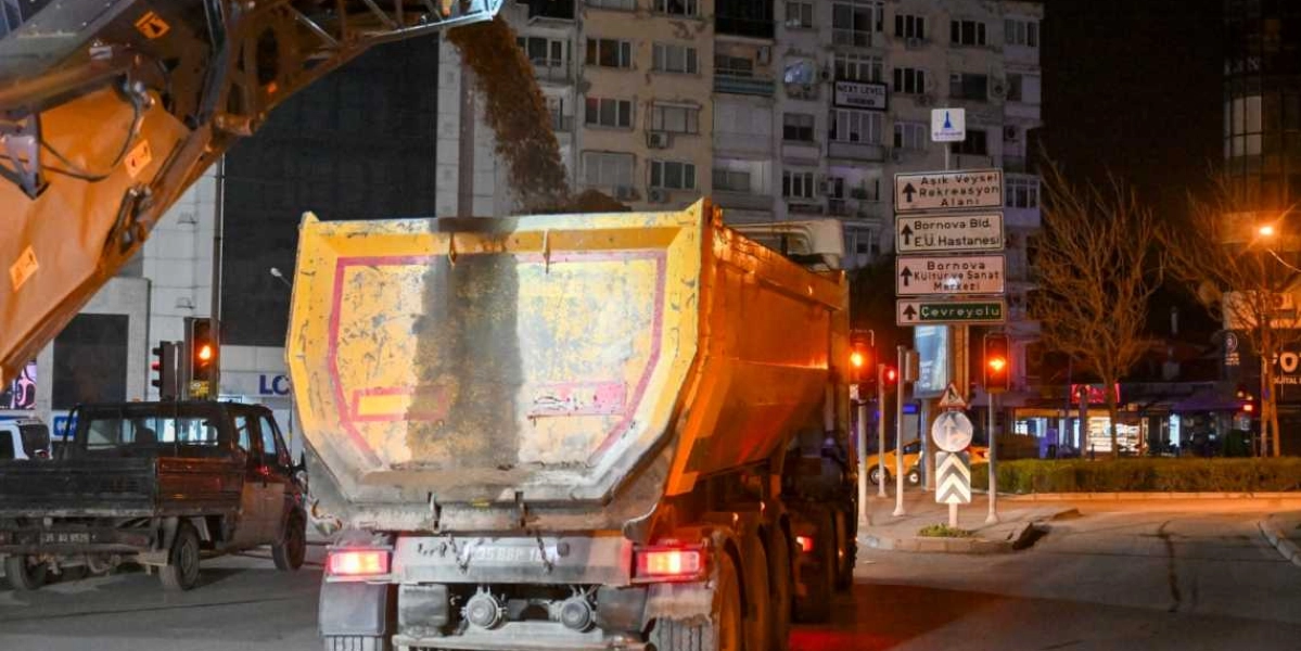 Gece saatlerinde Bornova'da bir iş makinesi, sarı bir damperli kamyonun kasasına asfalt malzemesi boşaltıyor. Arka planda çok katlı binalar ve üzerinde Aşık Veysel Rekreasyon Alanı, Ege Üniversitesi Hastanesi, Bornova Kültür ve Sanat Merkezi ile Çevreyolu yönlerini gösteren trafik tabelaları yer alıyor.