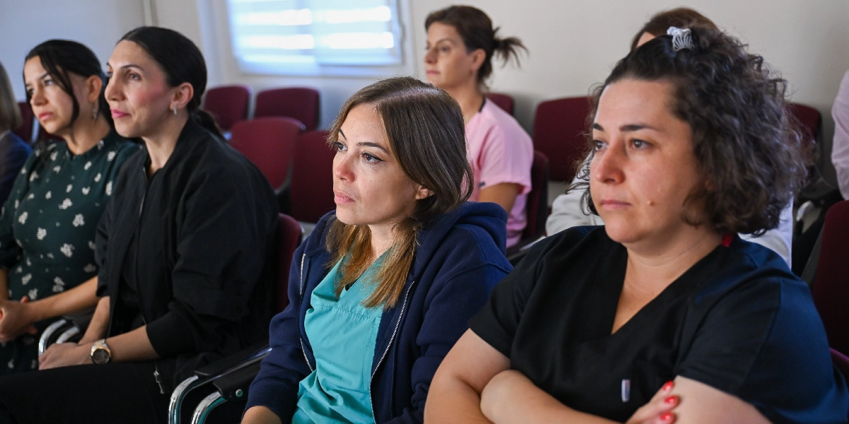 Bir konferans salonunda yan yana oturmuş, dikkatle dinleyen bir grup kadın. Ön planda, üzerinde lacivert bir hırka ve turkuaz bir üst bulunan bir kadın ile yanında siyah üniformalı, kolları kavuşturulmuş başka bir kadın yer alıyor. Arka planda ise koyu renkli kıyafetler giymiş diğer katılımcılar ve pembe bir üst giymiş bir sağlık çalışanı bulunuyor.