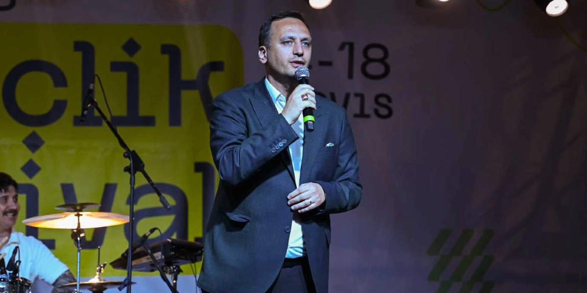 Koyu renk ceket ve beyaz gömlek giymiş bir erkek, sahnede elinde mikrofonla konuşma yapıyor. Arka planda üzerinde kısmen görünen 