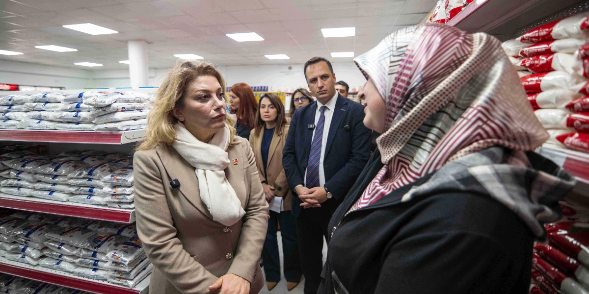 CHP Kadın Kolları Başkanı Asu Kaya, bir market reyonunun önünde başörtülü bir kadınla sohbet ederken, yanında Bornova Belediye Başkanı Ömer Eşki ve diğer yetkililer yer alıyor. Arka planda raflarda paketlenmiş gıda ürünleri dizili durumda.