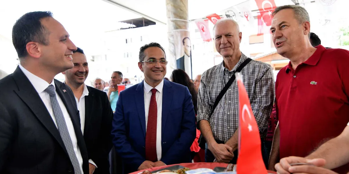 Bornova Belediye Başkanı Mustafa İduğ ve beraberindeki heyet, bir etkinlik sırasında sohbet ederken görülüyor. Ön planda, lacivert takım elbiseli ve kırmızı kravatlı Mustafa İduğ ile yanında kırmızı polo yaka tişörtlü bir kişi yer alıyor. Arka planda ise Atatürk posteri ve Türk bayrakları asılı olan açık hava alanında, takım elbiseli ve günlük kıyafetli diğer katılımcılar gülümseyerek birbirleriyle konuşuyor.
