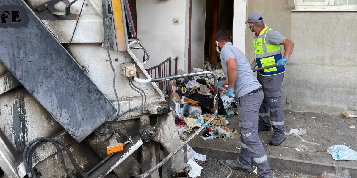 Bornova Belediyesi üniformalı iki temizlik görevlisi, bir apartman girişinde birikmiş olan yoğun çöp yığınını küreklerle temizleyerek hemen yanlarında duran çöp kamyonuna yüklüyor. Görevliler maske ve eldiven kullanarak çalışıyor.