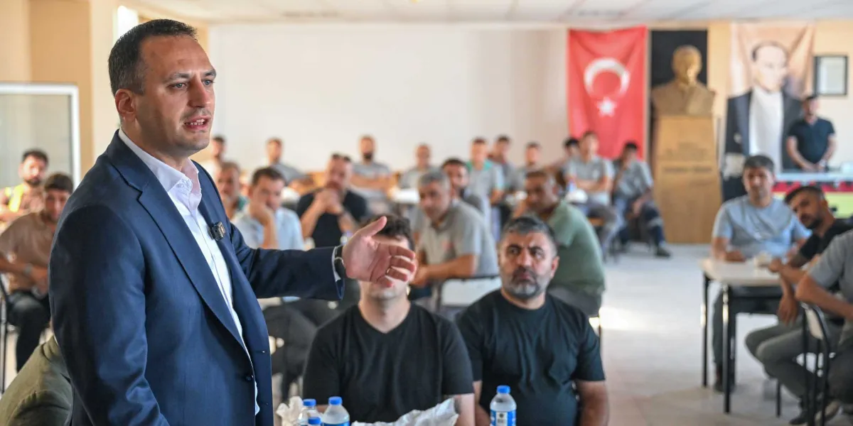 Koyu renk ceket ve beyaz gömlek giymiş bir erkek, kapalı bir toplantı salonunda oturan itfaiye personeline hitaben konuşma yapıyor. Arka planda duvarda asılı Türk bayrağı ve Mustafa Kemal Atatürk'ün portresi yer alıyor. İtfaiyeciler masalarda oturarak konuşmacıyı dinliyor.