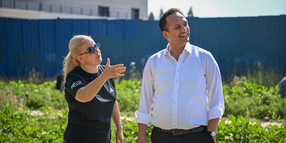 Siyah tişörtlü, sarı saçlı bir kadın, yanında duran beyaz gömlekli bir erkeğe eliyle bahçeyi işaret ederek bir şeyler anlatıyor. İkili, arkalarında mavi bir çit bulunan yeşil bitkilerle dolu bir alanda, güneşli bir havada sohbet ediyor.