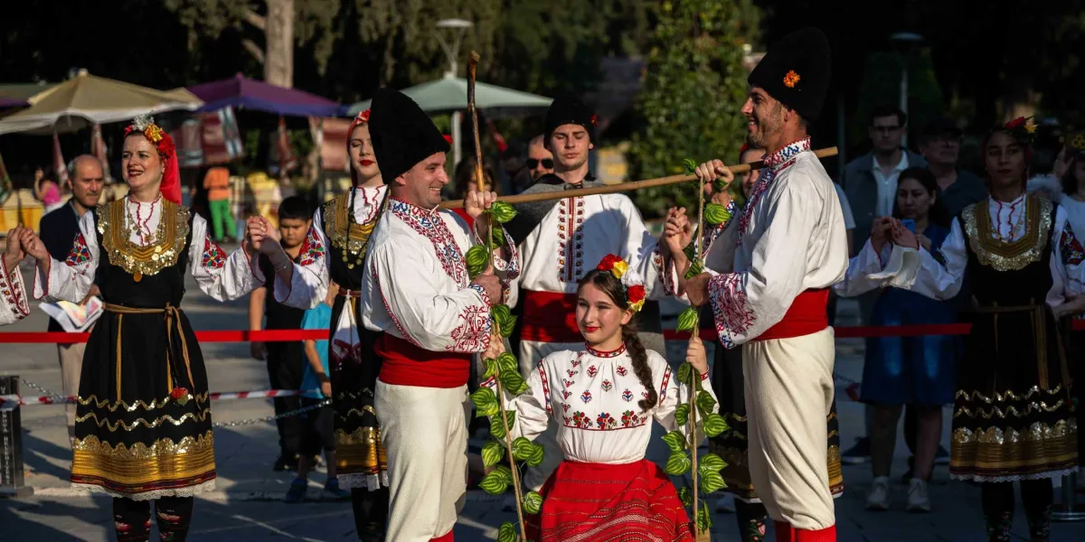 Geleneksel kıyafetler giymiş bir grup dansçı, açık havada el ele tutuşarak bir halk oyunu sergiliyor. Erkekler beyaz gömlek, kırmızı kuşak ve yüksek siyah başlıklar takarken, kadınlar işlemeli yelekler, altın rengi detaylı etekler ve çiçekli baş süsleri kullanıyor. Grubun merkezinde, iki erkek dansçının yatay bir sopa yardımıyla havada tuttuğu iplere tutunarak oturan genç bir kız bulunuyor. Arka planda ise etkinliği izleyen insanlar ve ağaçlıklı bir park alanı görülüyor.