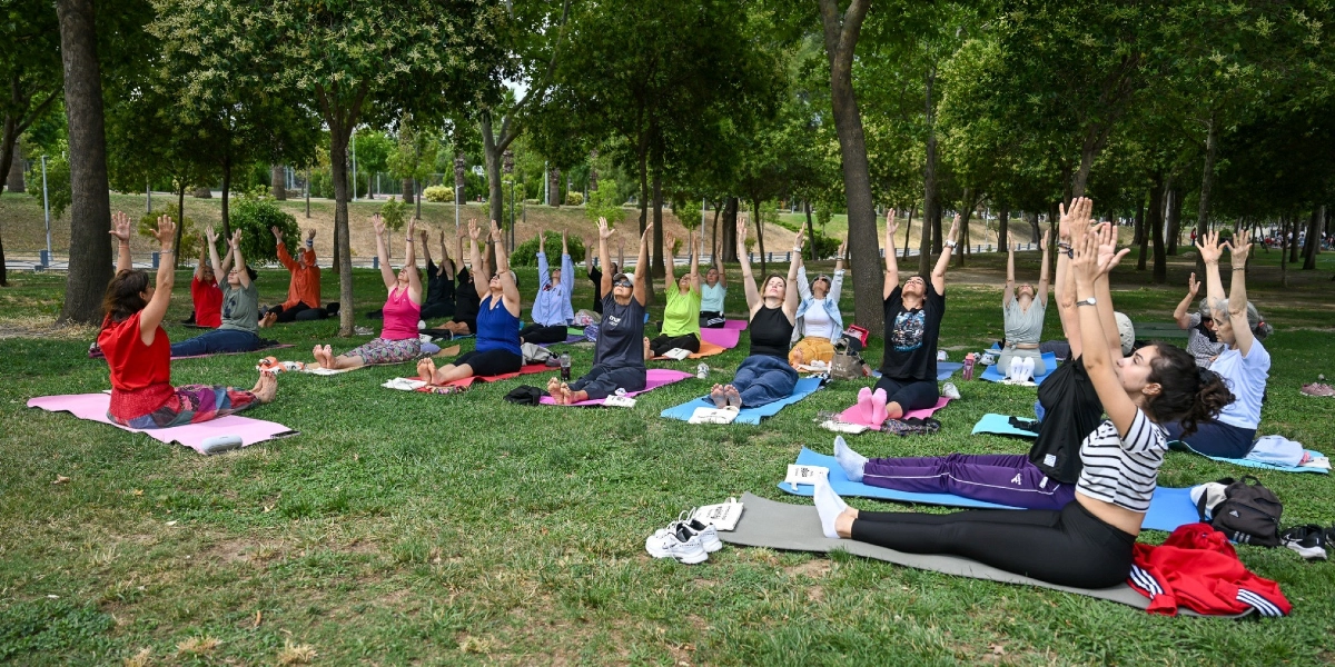 Ağaçlık bir park alanında, çimlerin üzerine serilmiş matlar üzerinde oturan ve ellerini havaya kaldırmış yoga yapan bir grup kadın.