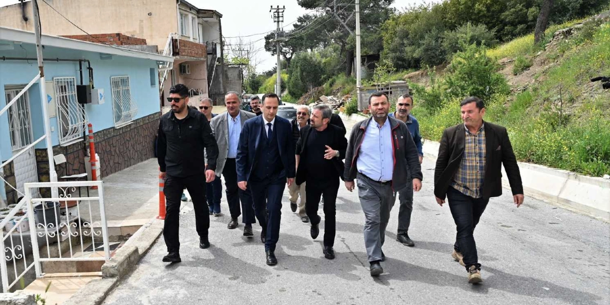 Belediye Başkanı Filiz Ceritoğlu Sengel'in de aralarında bulunduğu bir grup erkek, açık havada, asfalt bir yolda yan yana yürüyerek ilerliyor. Başkan, koyu renkli takım elbisesi ve kravatıyla grubun ön sıralarında yer alıyor. Yanındaki kişiler günlük kıyafetler, ceketler ve gömlekler giymiş durumda. Grubun sol tarafında açık mavi boyalı, pencerelerinde demir parmaklıklar bulunan tek katlı bir ev, sağ tarafında ise yeşilliklerle kaplı hafif eğimli bir arazi bulunuyor.