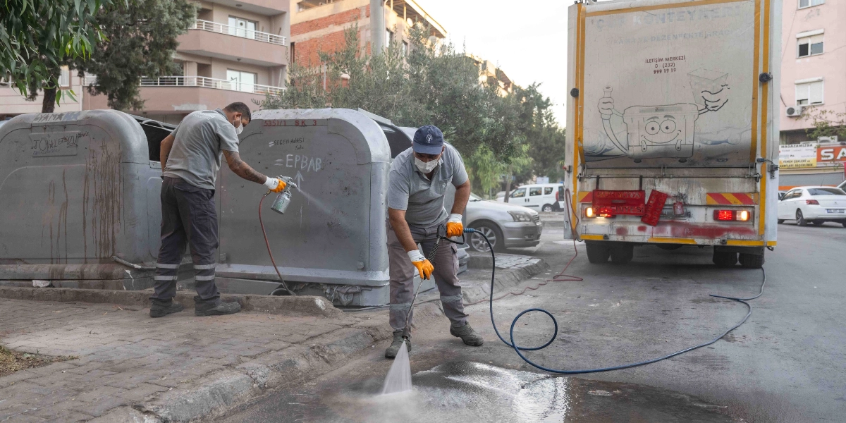 Belediye temizlik görevlileri, Bornova'da bir sokak üzerinde çöp konteynerlerini dezenfekte ediyor. Görevlilerden biri elindeki püskürtücüyle gri renkli metal çöp konteynerini temizlerken, diğeri basınçlı su hortumuyla konteynerin önündeki zemini yıkıyor. Arkalarında üzerinde 
