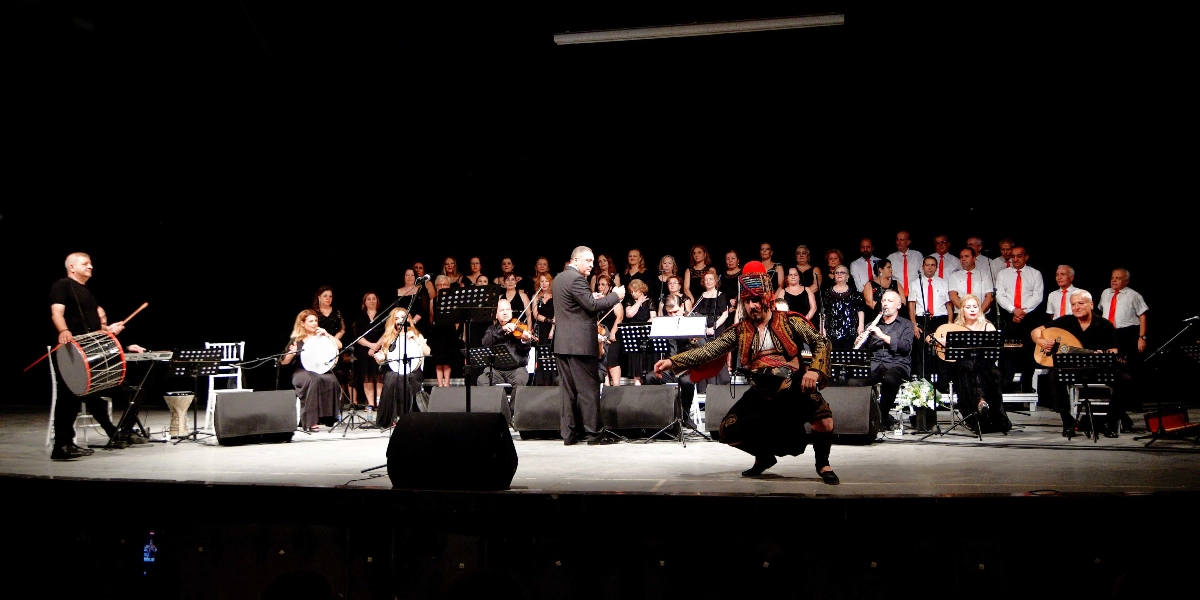 Siyah bir sahne üzerinde, orkestra şefinin yönettiği geniş bir koro ve müzisyen grubu performans sergiliyor. Sahnenin ön planında geleneksel kıyafetli bir zeybek dansçısı çömelmiş pozisyonda yer alırken, sol tarafta davul çalan bir müzisyen, sağ tarafta ise ud çalan bir sanatçı bulunuyor. Arka planda ise siyah kıyafetli kadınlar ve beyaz gömlekli, kırmızı kravatlı erkeklerden oluşan kalabalık bir koro grubu, nota sehpalarının önünde ayakta duruyor.