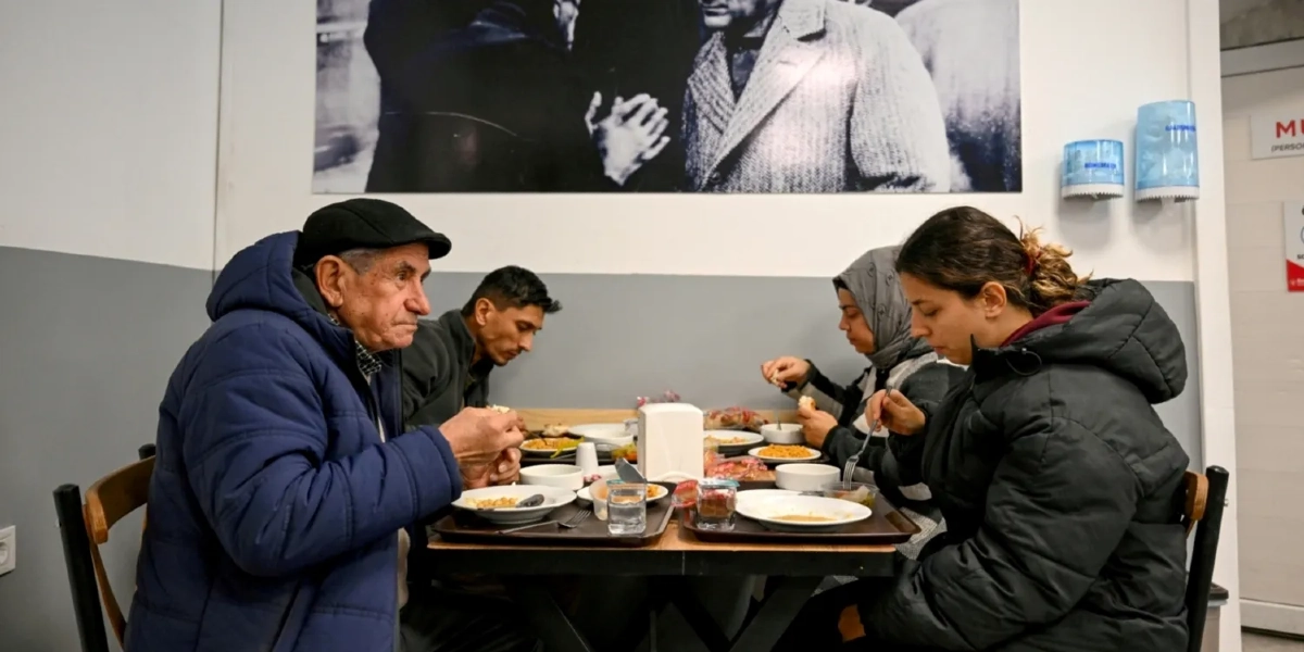 Bir yemekhanede masanın etrafında oturmuş yemek yiyen dört kişi; ön planda lacivert montlu ve kasketli yaşlı bir adam, yanında genç bir erkek, karşısında ise başörtülü bir kadın ve koyu renk montlu genç bir kadın bulunuyor. Masanın üzerinde tabaklar, bardaklar ve peçetelik yer alıyor. Arka duvarda ise siyah beyaz bir Atatürk fotoğrafı asılı.