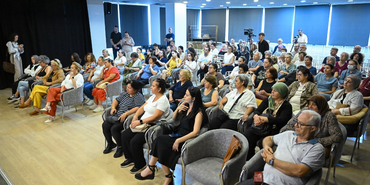 Modern bir konferans salonunda, ağırlıklı olarak kadınlardan oluşan kalabalık bir dinleyici grubu gri koltuklarda oturarak bir sunumu takip ediyor. Salonun ön tarafında, sahneye dönük şekilde dizilmiş çok sayıda katılımcı dikkatle dinlerken, arka planda profesyonel bir kamera ve çekim yapan bir görevli yer alıyor. Salonun genelinde aydınlık bir atmosfer hakim ve katılımcılar günlük kıyafetlerle oturuyor.
