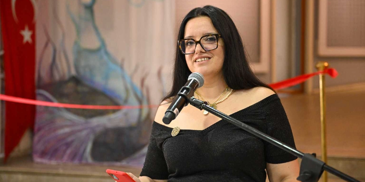 Siyah, omuzları açık bir bluz giymiş, uzun siyah saçlı ve gözlüklü bir kadın, mikrofon başında konuşma yapıyor. Kadının arkasında, üzerinde deniz kızı figürü bulunan bir pano ve sol tarafta asılı duran bir Türk bayrağı yer alıyor. Ön planda ise açılış törenlerinde kullanılan kırmızı bir kurdele görülüyor.