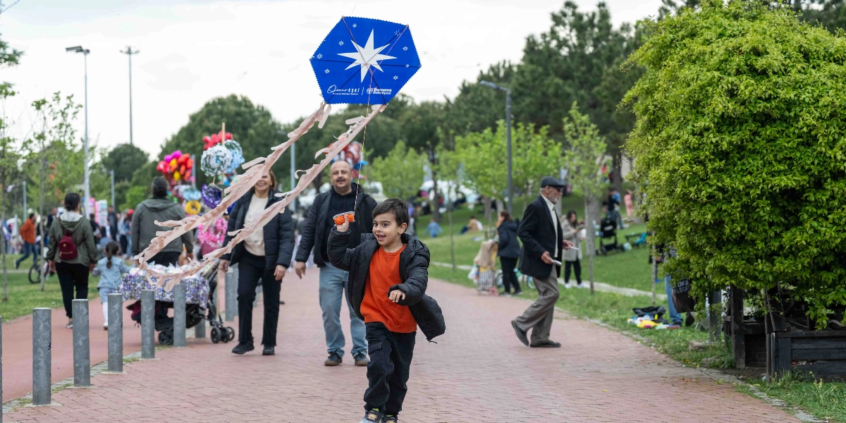 Açık havada, park yolunda koşan küçük bir çocuk, elinde üzerinde Bornova Belediyesi logosu ve beyaz bir yıldız bulunan altıgen şeklindeki mavi uçurtmayı uçuruyor. Çocuğun arkasında yürüyen yetişkinler ve çevrede vakit geçiren insanlar bulunuyor. Arka planda yeşil ağaçlar ve parkın genel atmosferi görülüyor.