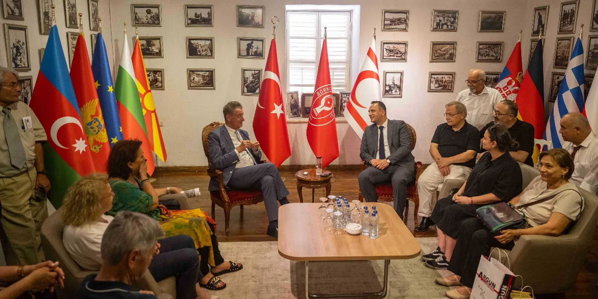 İç mekanda, duvarları siyah beyaz tarihi fotoğraflarla kaplı bir odada, çeşitli ülkelerin bayraklarının önünde bir araya gelmiş bir grup insan oturuyor. Ortada karşılıklı koltuklarda oturan iki erkek ve onları çevreleyen katılımcılar, resmi bir toplantı veya sohbet ortamında bulunuyor. Arka planda Türkiye Cumhuriyeti bayrağı, Kuzey Kıbrıs Türk Cumhuriyeti bayrağı ve Bornova Belediyesi'ne ait olduğu anlaşılan logolu bir flama yer alıyor. Odanın genelinde diplomatik veya kültürel bir buluşma atmosferi hakim.