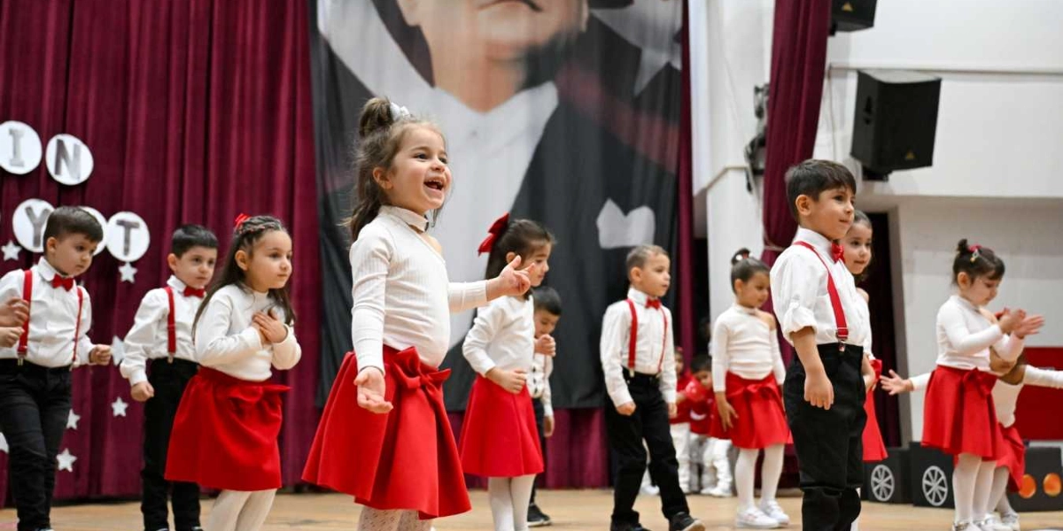 Beyaz gömlek, kırmızı papyon ve kırmızı askılı pantolon giyen anaokulu öğrencileri, arkalarında büyük bir Atatürk posterinin bulunduğu sahnede Cumhuriyet Bayramı kutlamaları kapsamında dans ediyor. Çocuklar neşeli bir ifadeyle el çırpıp hareket ederken, kız öğrenciler beyaz üst ve kırmızı etek, erkek öğrenciler ise beyaz gömlek ve siyah pantolon giyiyor.