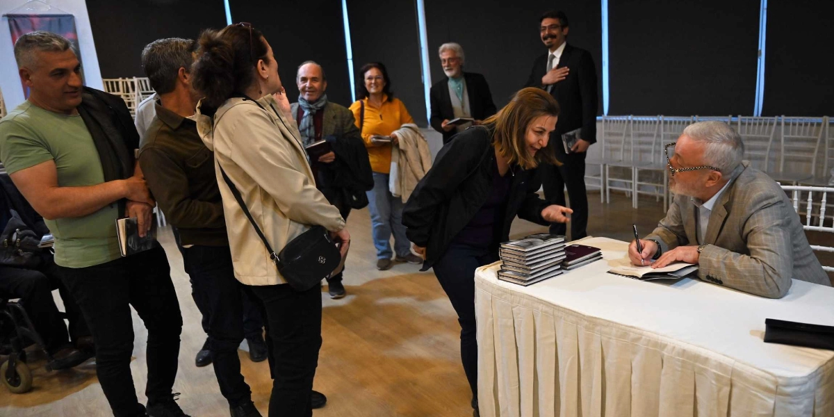İç mekanda düzenlenen bir imza gününde, gri ceketli ve gözlüklü bir yazar masasında oturmuş kitap imzalamaktadır. Yazarın önündeki masada bir yığın kitap dururken, önünde sıraya girmiş katılımcılar yazarla sohbet etmekte ve imzalı kitaplarını beklemektedir. Arka planda ise etkinliği izleyen diğer katılımcılar ayakta durmaktadır.