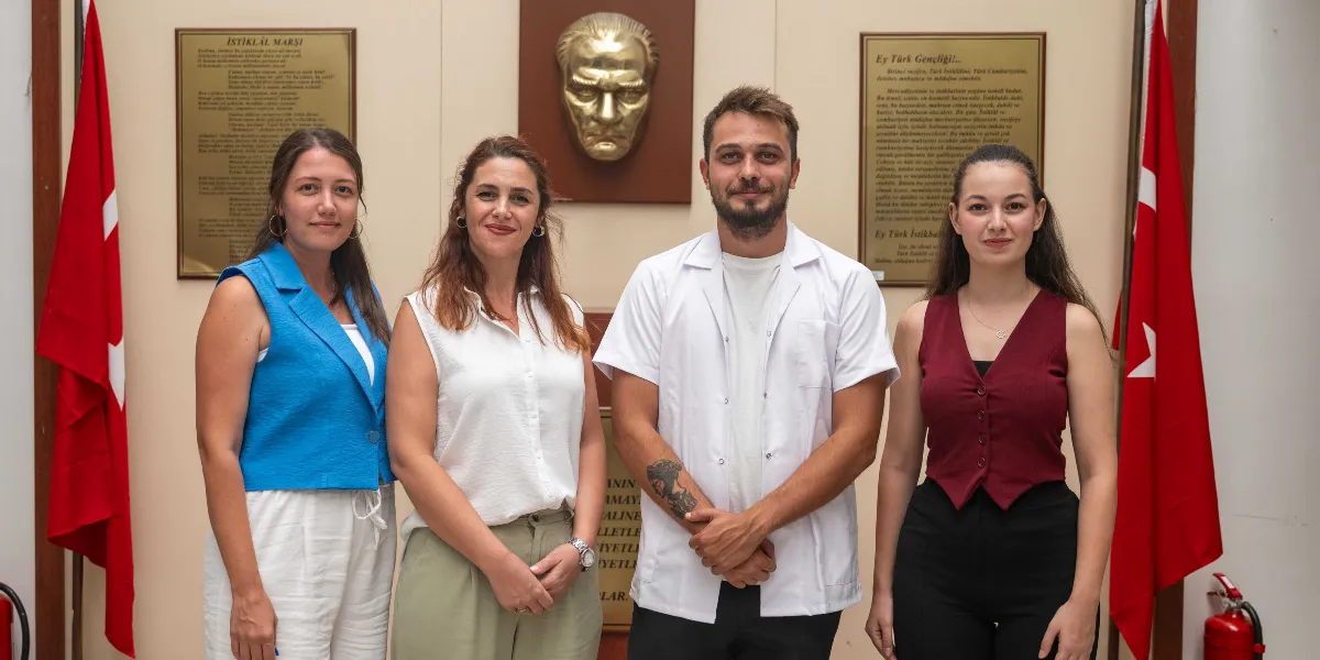 Arka planda duvarda asılı Atatürk kabartması, İstiklal Marşı ve Gençliğe Hitabe metinlerinin bulunduğu, iki yanında Türk bayraklarının yer aldığı bir odada, yan yana duran iki kadın ve bir erkekten oluşan üç kişilik grup. Soldaki kadın mavi yelek ve beyaz pantolon, ortadaki kadın beyaz kolsuz bluz ve bej pantolon, sağdaki erkek ise beyaz kısa kollu bir önlük giymiş şekilde kameraya gülümseyerek bakıyor.