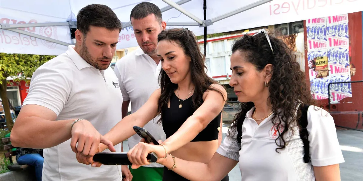 Bornova Belediyesi logolu beyaz tişört giyen iki görevli, siyah spor kıyafetli bir kadınla birlikte dış mekanda bir stant önünde duruyor. Görevlilerden biri elindeki cihazı işaret ederken, diğer görevli ve kadın cihazı birlikte tutuyor. Arka planda üzerinde 