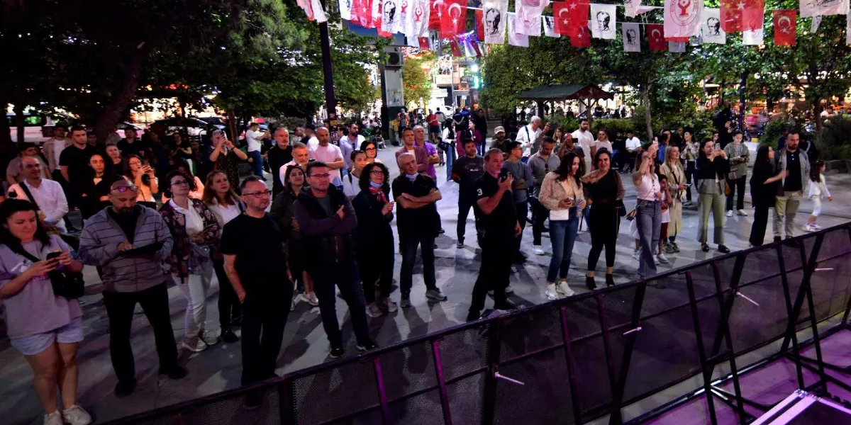 Akşam saatlerinde bir meydanda toplanmış, sahneye doğru bakan kalabalık bir grup insan. Meydanın üzeri, üzerinde Türk bayrakları ve Atatürk görselleri bulunan flamalarla süslenmiş. İnsanlar ayakta duruyor, bazıları ellerindeki telefonlarla çekim yapıyor veya sahneyi izliyor. Ön planda metal bir bariyer hattı yer alıyor.